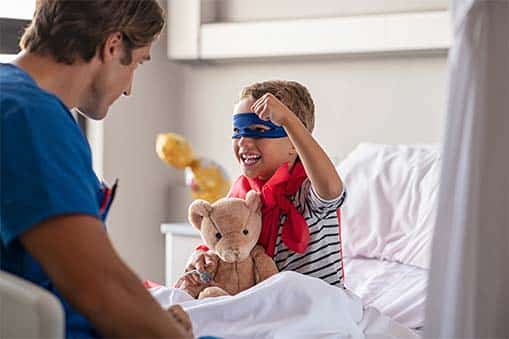 Little boy being a super hero in the hospital holding a teddy bear talking to a male nurse